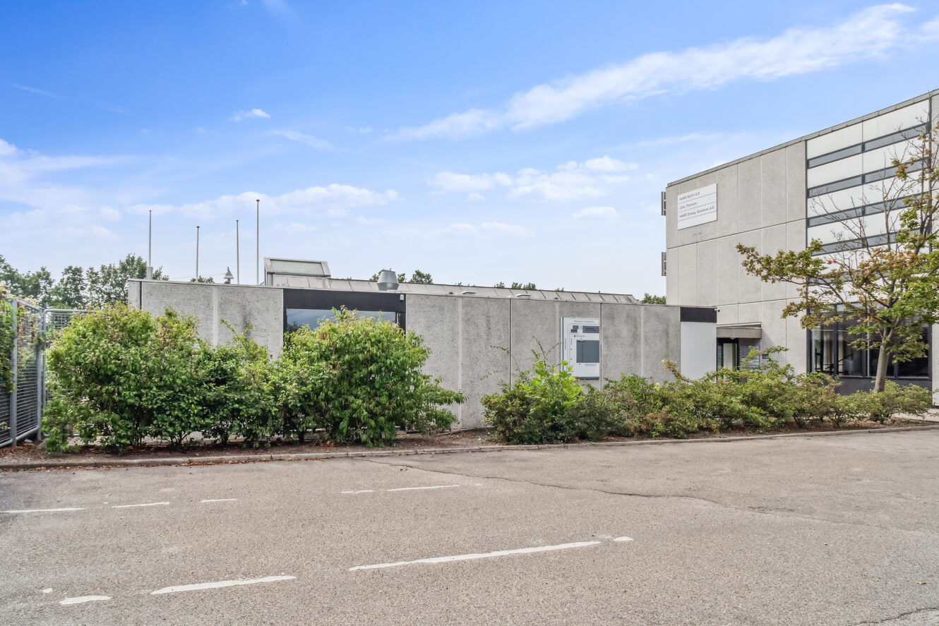 En erhvervsbygning i beton med store vinduer, omgivet af buske og en tom parkeringsplads under en delvis overskyet himmel.
