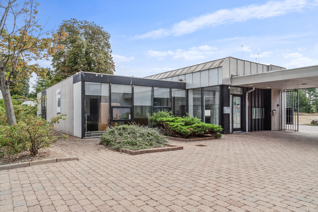 Erhvervsbygning i én etage med store glasvinduer, fladt tag og murstensbelagt indgangsparti, omgivet af grønt under en delvis overskyet himmel.