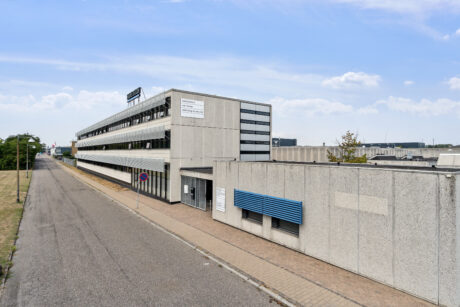 En lang, treetagers kontorbygning i beton med et skilt på taget, der støder op til en stille gade under en blå himmel.