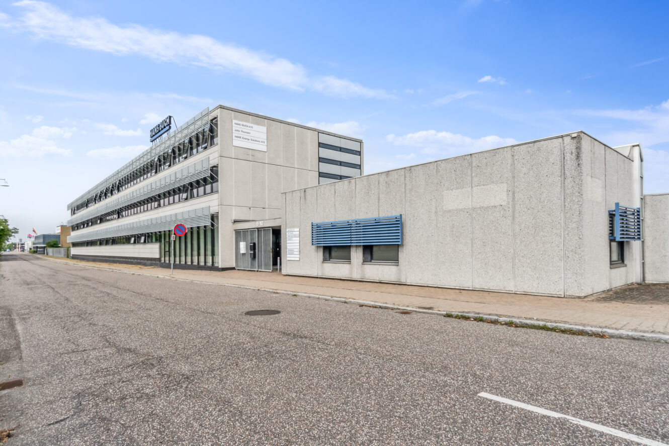 En moderne kontorbygning i tre etager med fladt tag og blå vinduesskærme ligger på en tom gade under en delvis overskyet himmel.