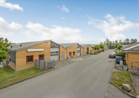 En række industrielle lagerbygninger med gule murstensvægge og grå tage ligger langs en asfalteret gade med parkerede biler under en delvist overskyet himmel.