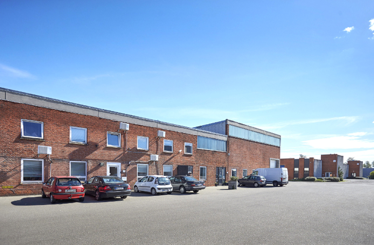 En forretningsbygning i mursten med flere biler og en varevogn parkeret foran, under en klar blå himmel.