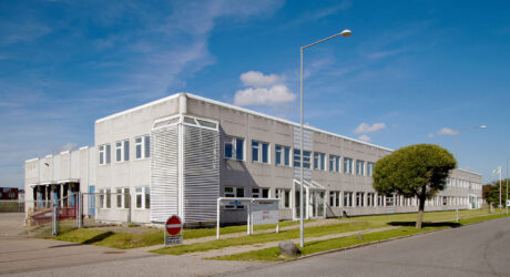 En moderne kontorbygning i to etager med hvid facade, store vinduer og skilte ved indgangen, som ligger på en stille gade under en blå himmel.