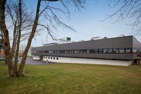 En moderne industri- eller kontorbygning i to etager med en mørk øvre facade og en hvid nedre etage, der støder op til et græsareal og træer med en parkeringsplads i baggrunden.