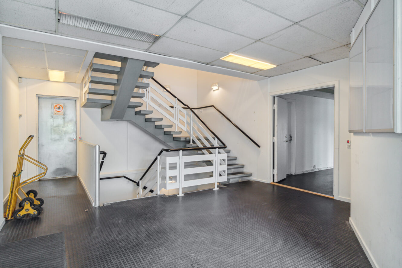 Bygningsinteriør i industristil med metaltrapper, gelændere, en dør til venstre og en gul sækkevogn på et sort struktureret gulv. Fluorescerende belysning over hovedet.