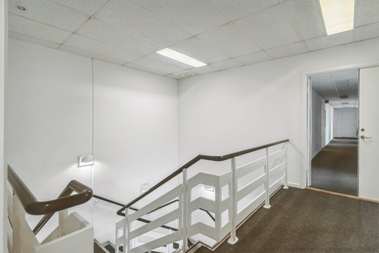 En hvid trappe med metalgelænder og en brun håndliste fører ned fra en gang med gulvtæppe i en kontorbygning med fluorescerende loftslamper.