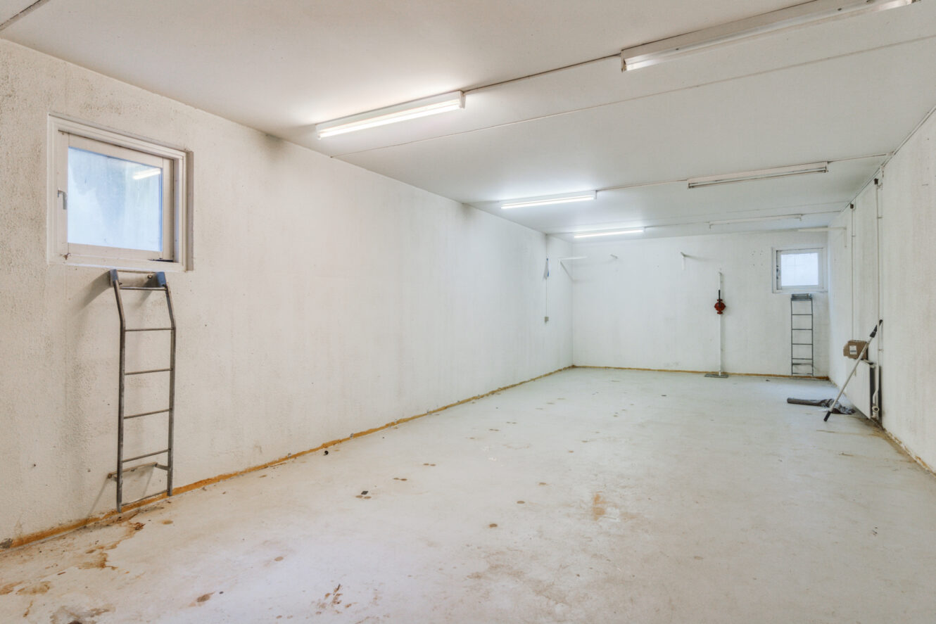 Tom, veloplyst garage med hvide vægge, to stiger, et vindue til venstre og spredt vedligeholdelsesværktøj langs den højre væg.