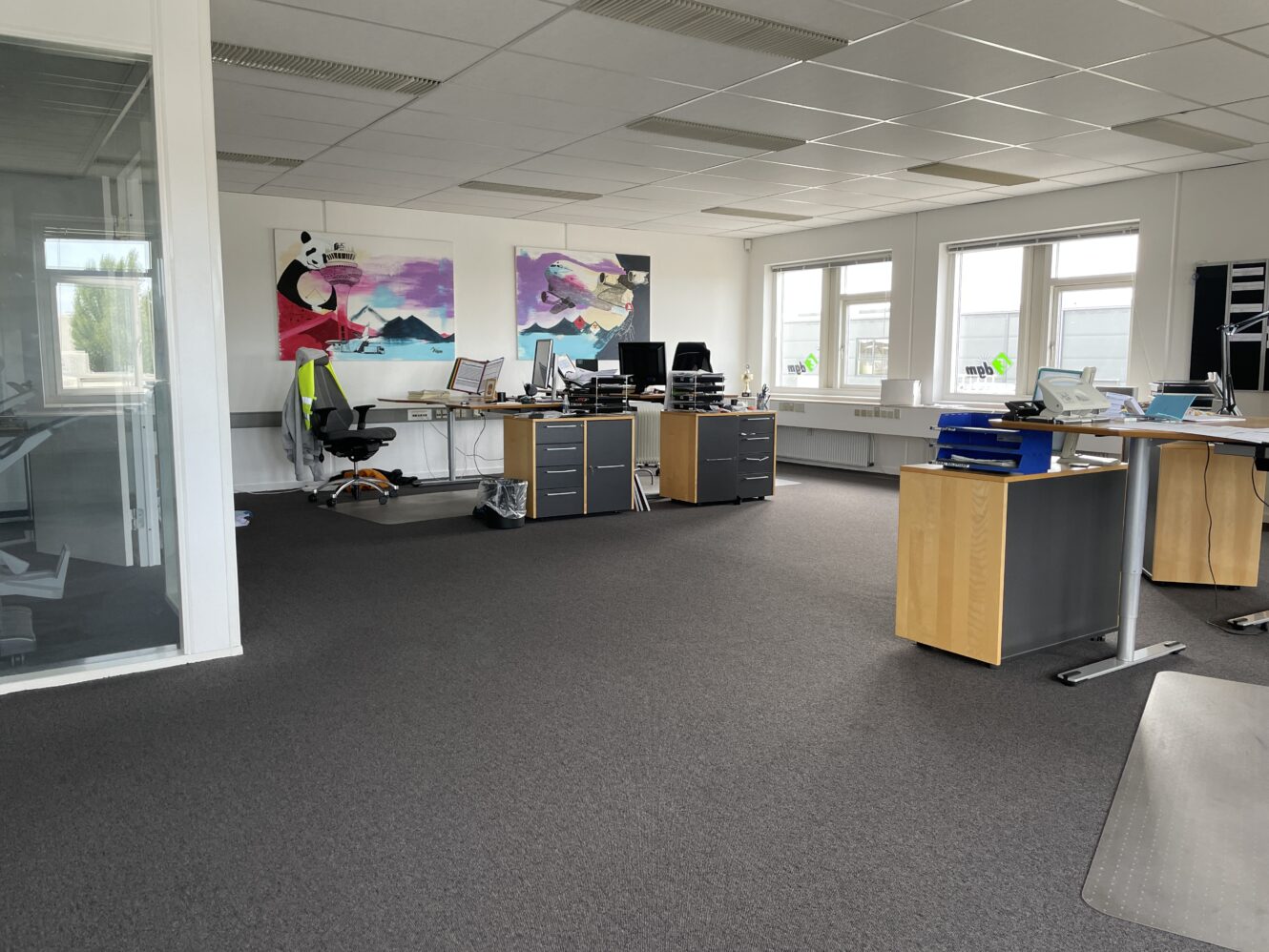 Moderne kontor med skriveborde, computere og kontorstole; kunstværker på væggen; store vinduer giver naturligt lys; nogle papirer og forsyninger på skrivebordene.