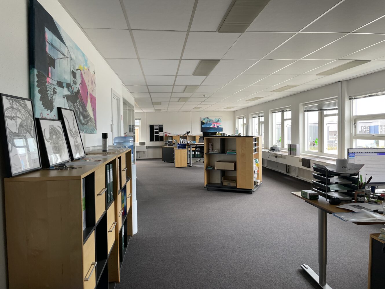 Åbent kontor med skriveborde, hylder, computere og store vinduer langs den ene væg. Indrammede kunstværker og dokumenter er udstillet på skabe. Rummet er tomt for mennesker.
