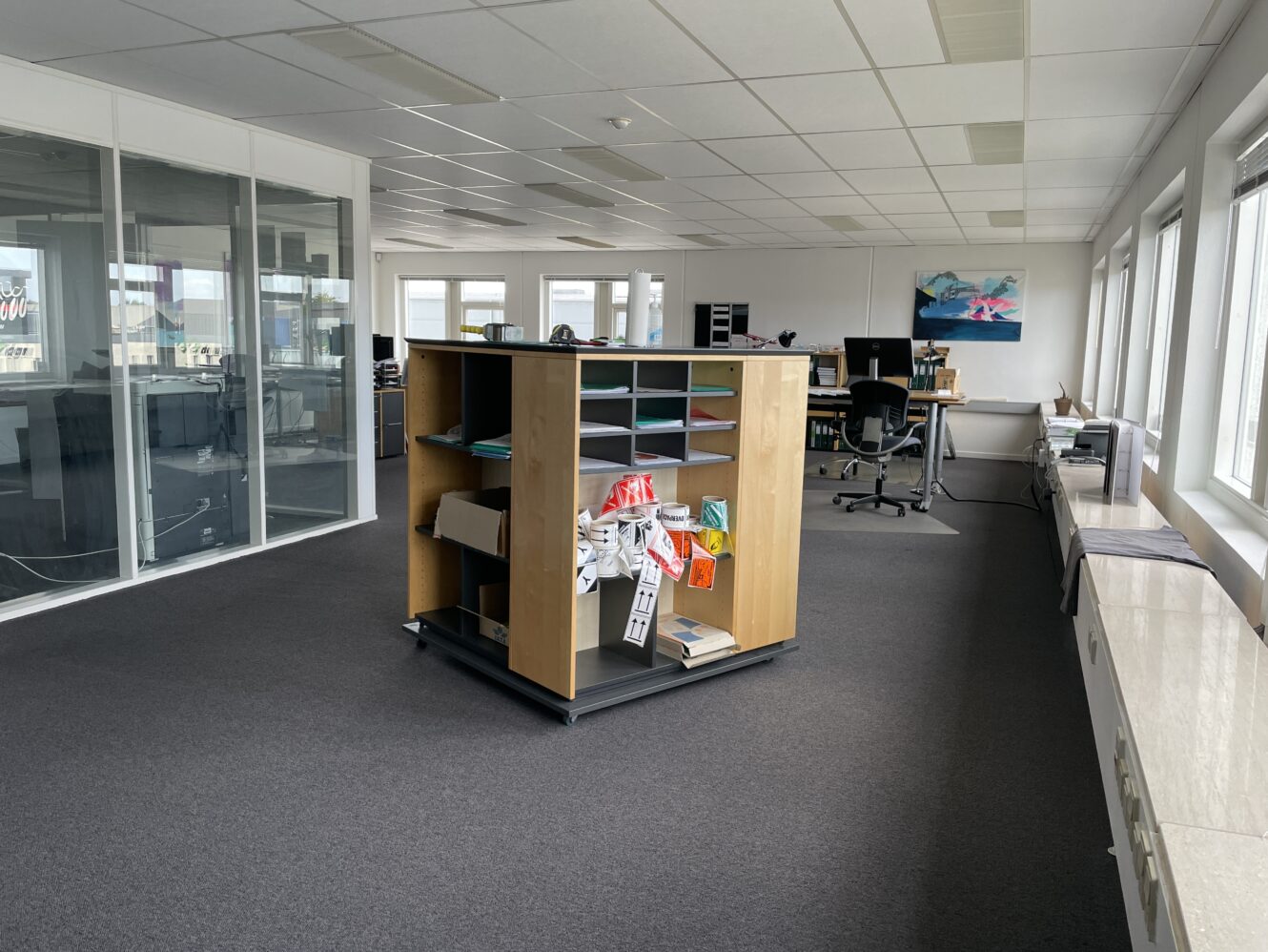 Et åbent kontorlandskab med skriveborde, stole, store vinduer og en central træreol med papirer og kontorartikler.