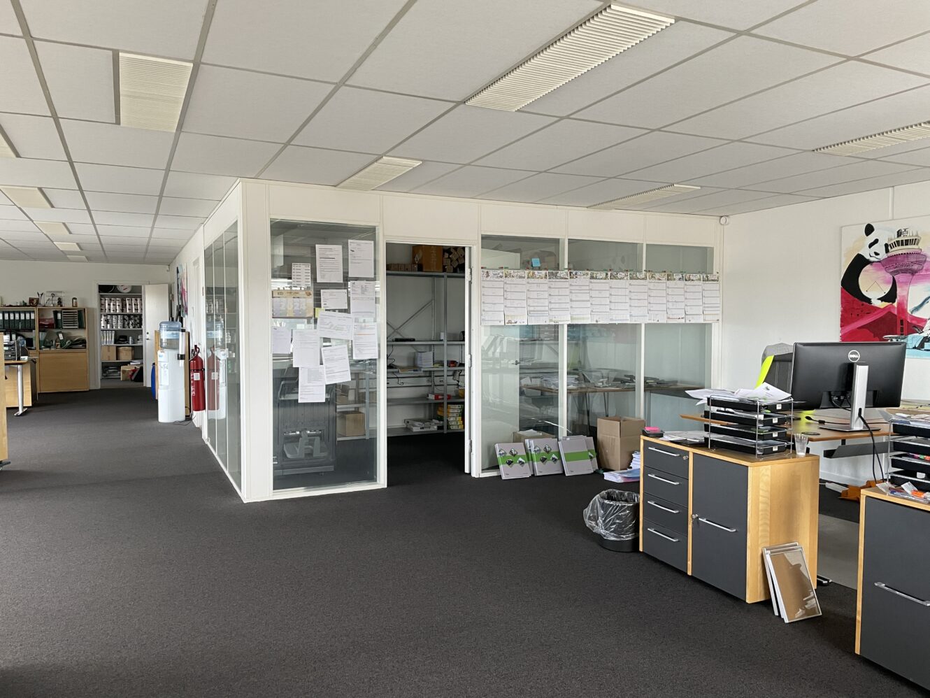 Moderne kontorlokaler med mødelokaler med glasvægge, skriveborde med kontorartikler, hylder og dokumenter, der er hængt op på glas; en pandaplakat på væggen.
