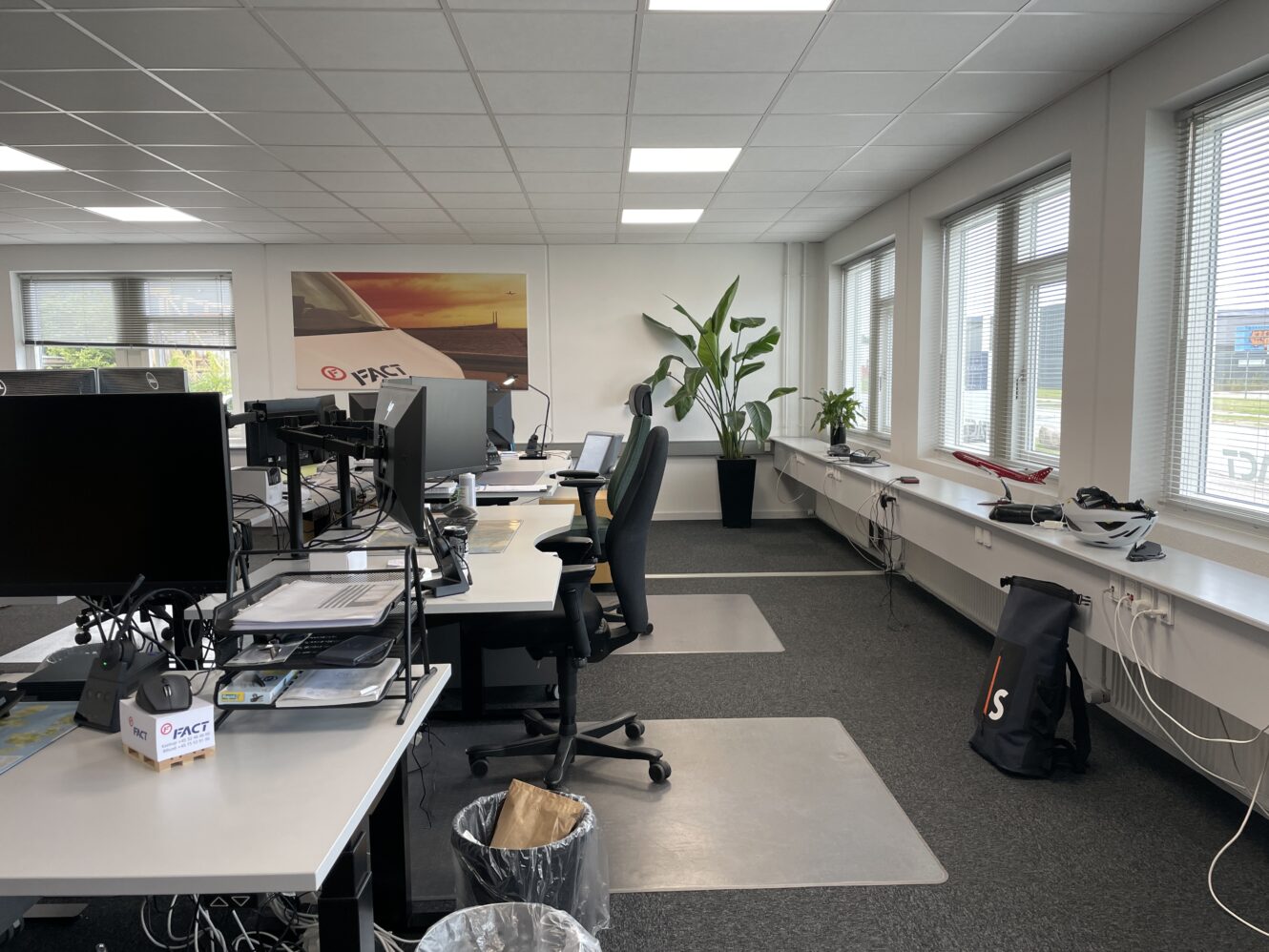 Moderne kontor med flere skriveborde, computere, kontorstole, en stor plante og vinduer, der lukker naturligt lys ind; vægkunst og modelfly dekorerer rummet.