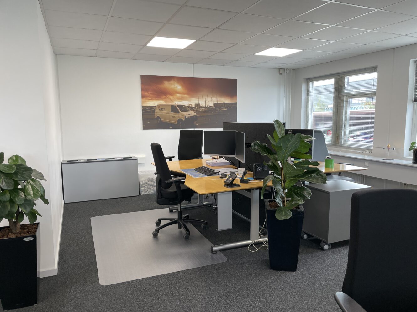 Et moderne kontor med to skriveborde, computerskærme, kontorstole, potteplanter og et stort vægbillede af lastbiler; vinduer lukker naturligt lys ind.