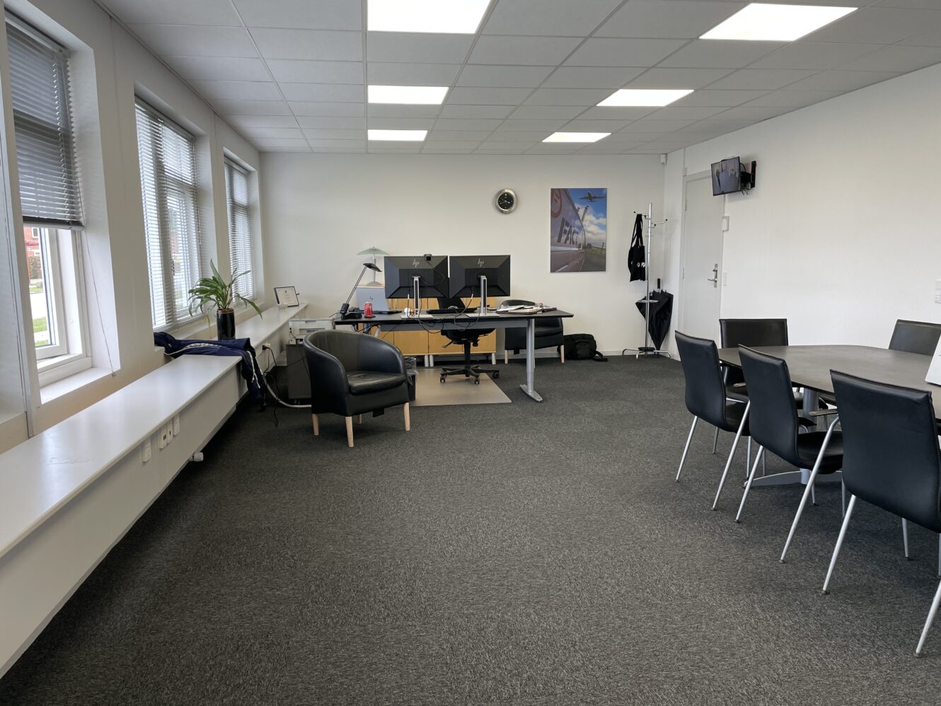 Moderne kontorlokaler med skriveborde, computerskærme, stole, et lille rundt mødebord, vægkunst, knagerække og vinduer, der lukker naturligt lys ind.