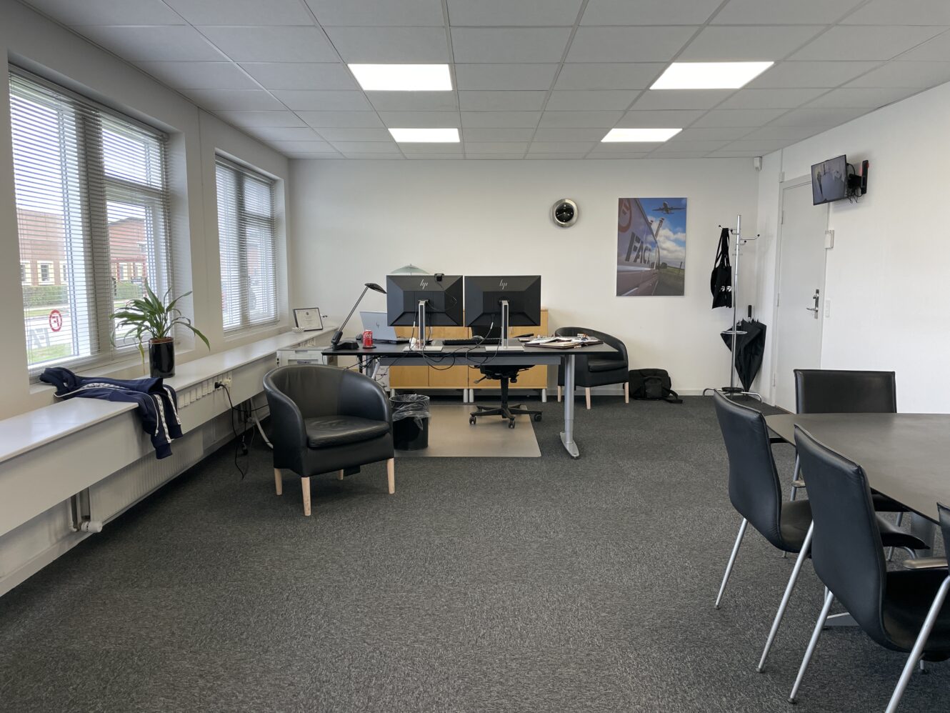 Et moderne kontor med skriveborde, computere, stole, et bord med fire stole, vægkunst og store vinduer, der lukker naturligt lys ind.