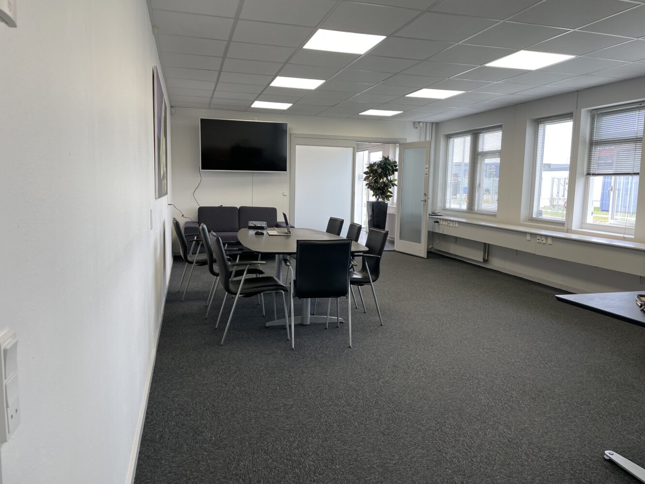 Et moderne konferencelokale med et rundt bord, flere stole, en stor væghængt skærm og vinduer langs den ene side, der lukker naturligt lys ind.