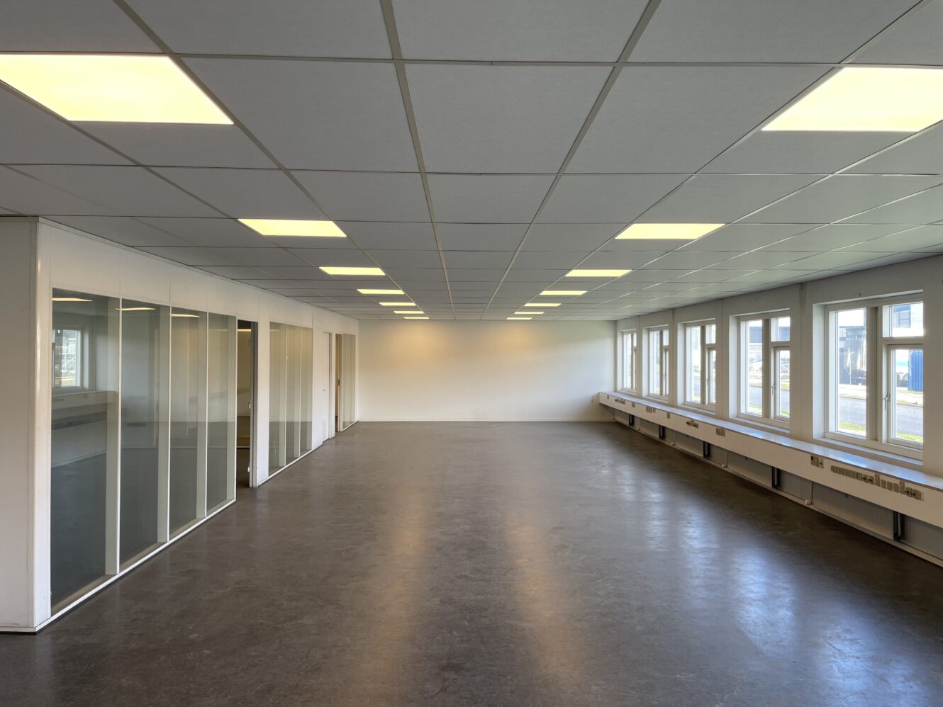 Tomt kontorlokale med mørkt gulv, glasvægge til venstre, store vinduer til højre og loftslamper med jævn afstand.