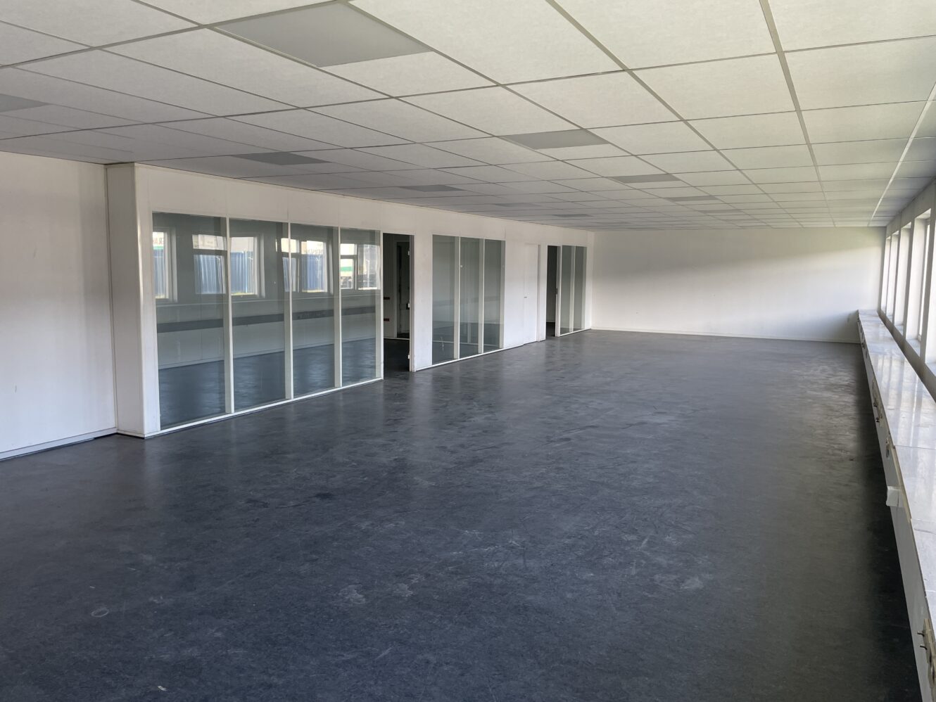 Tomt kontorlokale med store vinduer, glasvægge til venstre, hvide vægge og et mørkt gulv. Loftet har indbyggede rektangulære lyspaneler.