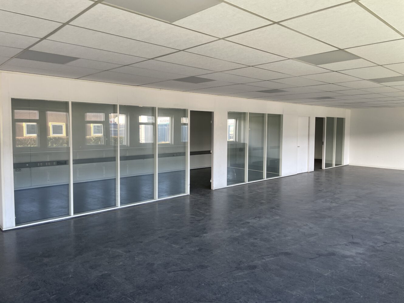 Tomme kontorlokaler med glasvægge, hvide loftspaneler og grå gulve; vinduerne afspejler bygningerne udenfor.