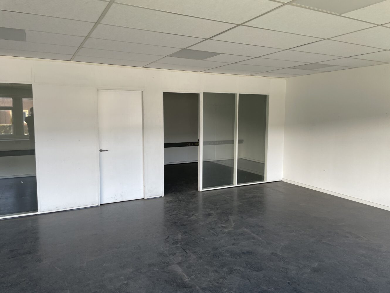 Tomt kontorlokale med hvide vægge, en lukket hvid dør og en glasskillevæg, der adskiller et tilstødende rum; mørkt gulv og et gittermønstret loft.