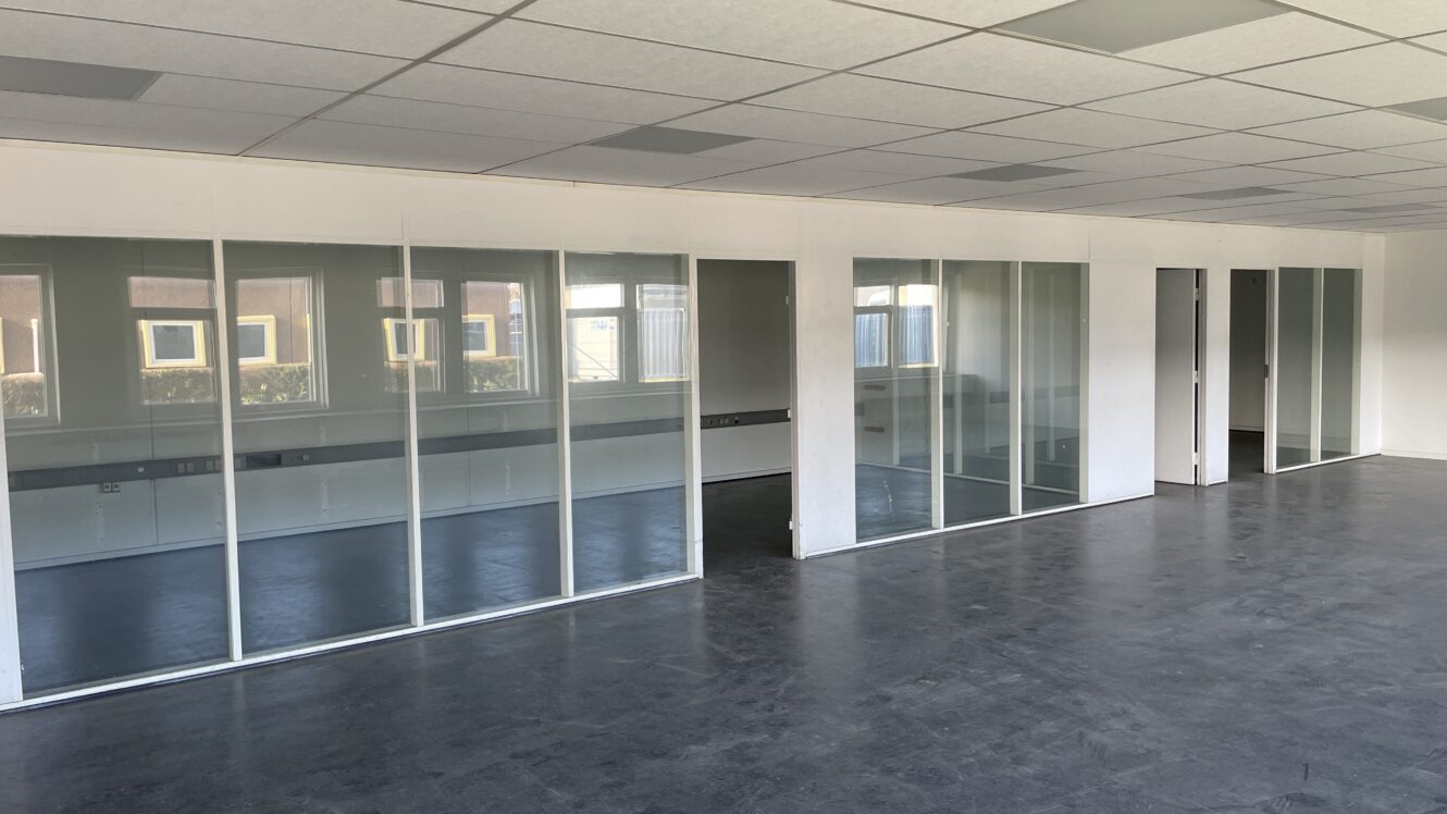 Tomt kontorlokale med glasskillevægge, mørkt gulv og et nedhængt loft med lysstofrør.