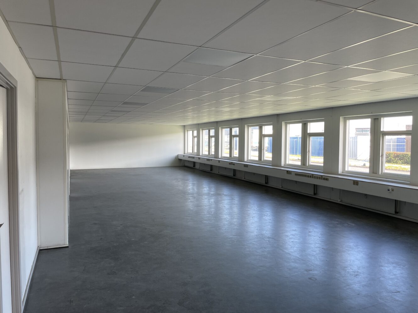 Et tomt, umøbleret rum med flisebelagt loft, gråt gulv, hvide vægge og store vinduer langs den ene side, der lukker naturligt lys ind.