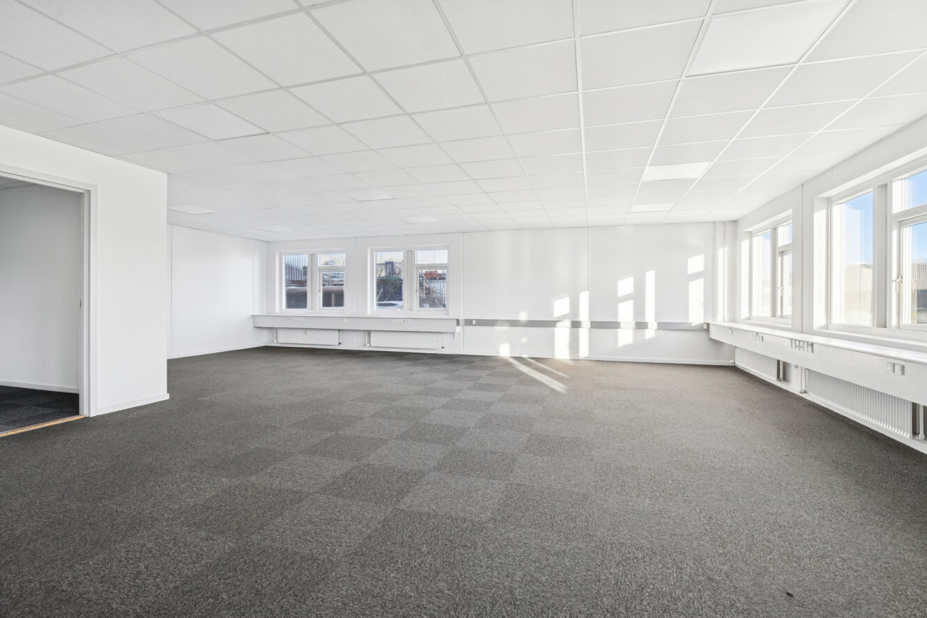 Tomt kontorlokale med store vinduer, gråt gulvtæppe, hvide vægge og et gittermønstret loft. Sollyset strømmer ind gennem vinduerne i højre side af lokalet.