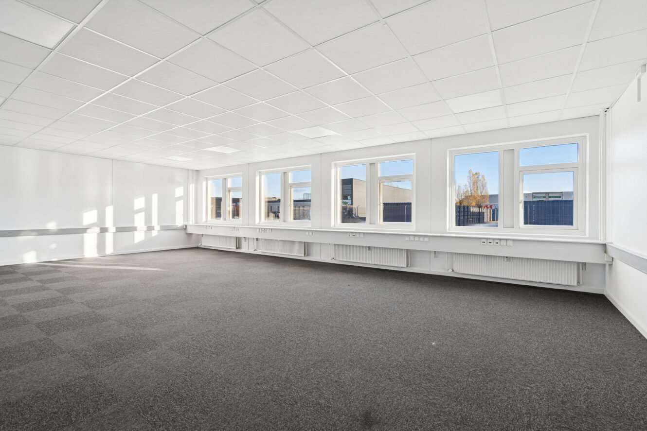 Tomt kontorlokale med gråt gulvtæppe, hvide vægge og flere store vinduer, der giver naturligt lys ind i lokalet.