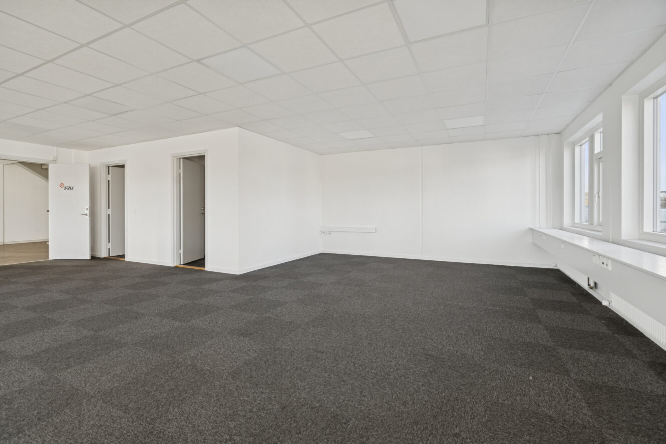 Stort tomt kontorlokale med gråt gulvtæppe, hvide vægge, nedhængte loftsplader og flere vinduer langs den ene side. Tre lukkede døre er synlige i baggrunden.