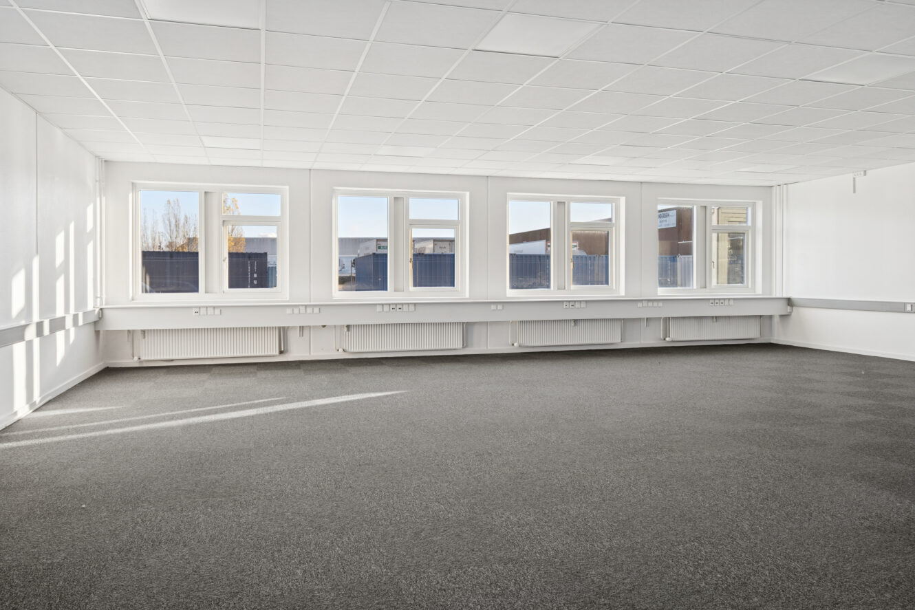 Tomt kontorlokale med gråt gulvtæppe, hvide vægge, hvidt loft og fem store vinduer, der lukker naturligt lys ind.