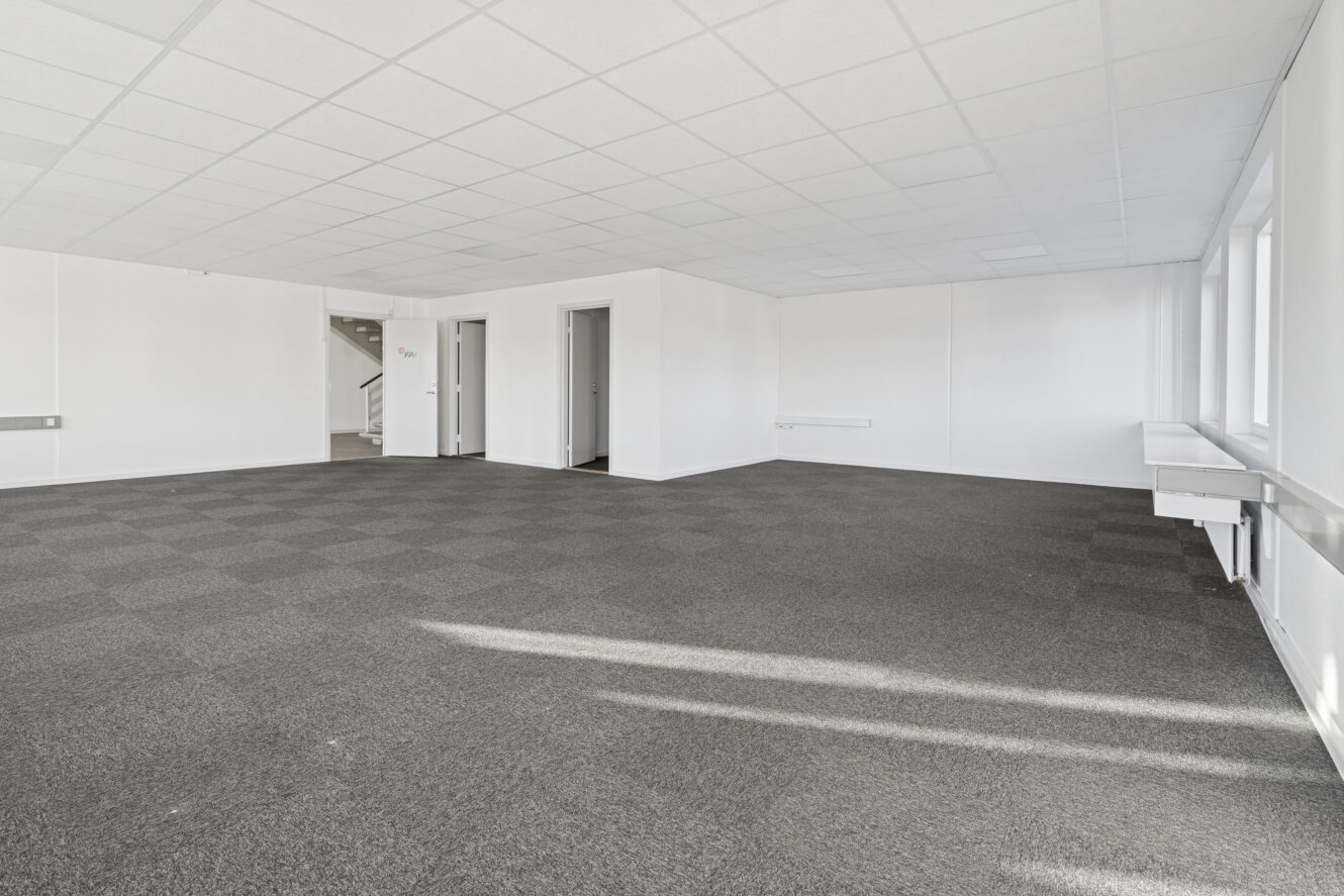 Tomt kontorlokale med gulvtæppe, hvide vægge, nedhængt loft, vinduer til højre og døråbninger på bagsiden.