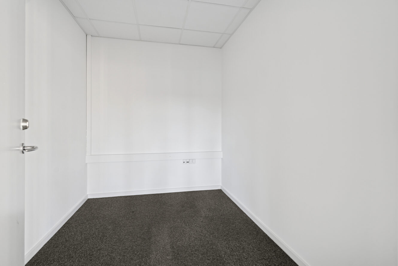 Et lille, tomt rum med hvide vægge, en lukket dør, et gråt gulvtæppe og et hvidt loft med paneler.