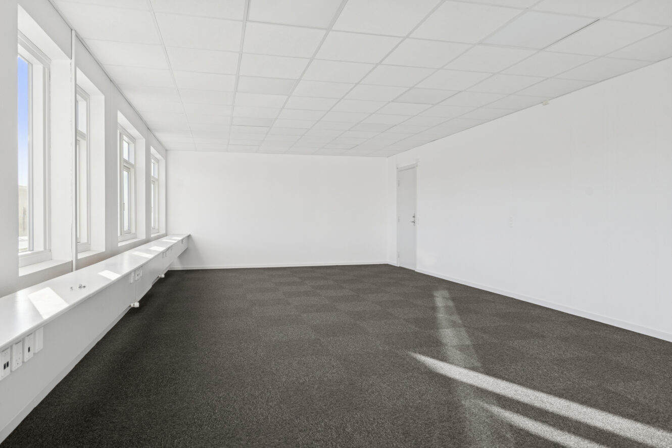 Et lyst, tomt kontorlokale med hvide vægge, et gråt gulvtæppe, flere vinduer langs den ene side og en lukket hvid dør i den anden ende.