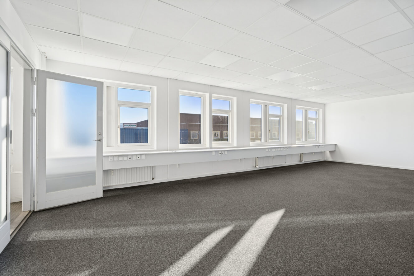 Tomt kontorlokale med gråt gulvtæppe, hvide vægge, en række vinduer, der lukker naturligt lys ind, og en glasdør til venstre.