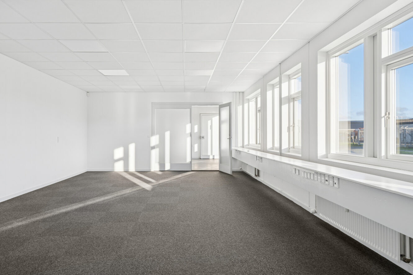 Tomt kontorlokale med hvide vægge, store vinduer til højre, der lukker sollys ind, gråt gulvtæppe og et hvidt loft med indbyggede lamper.