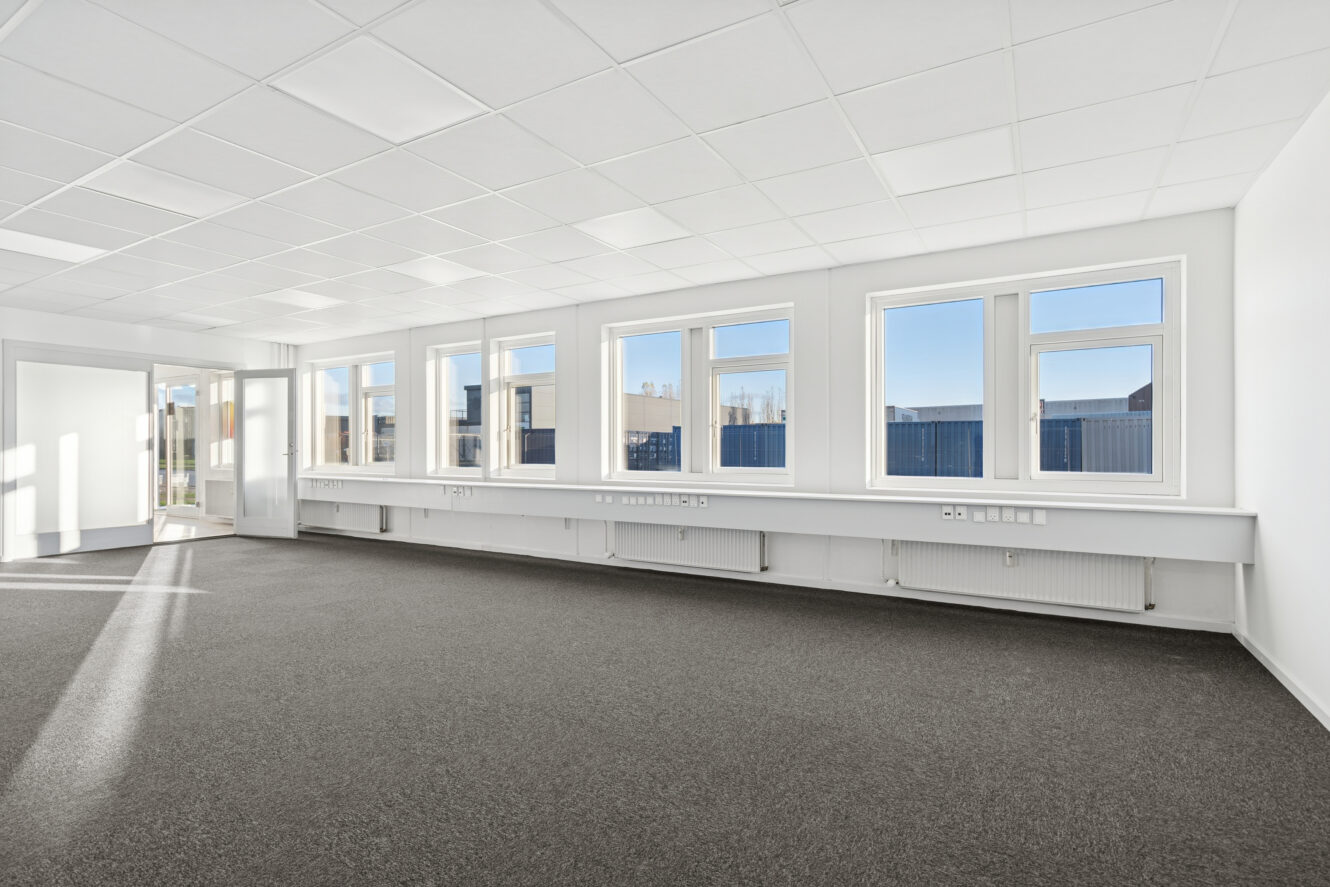 Et stort, tomt kontorlokale med hvide vægge, flere vinduer, der lukker naturligt lys ind, og et gråt gulvtæppe.
