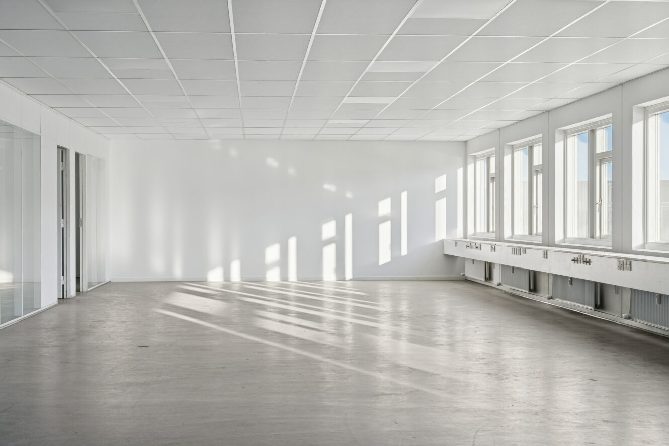 Tomt, moderne kontorlokale med store vinduer til højre, hvide vægge, loftplader og bart gulv; sollyset kaster skygger hen over lokalet.