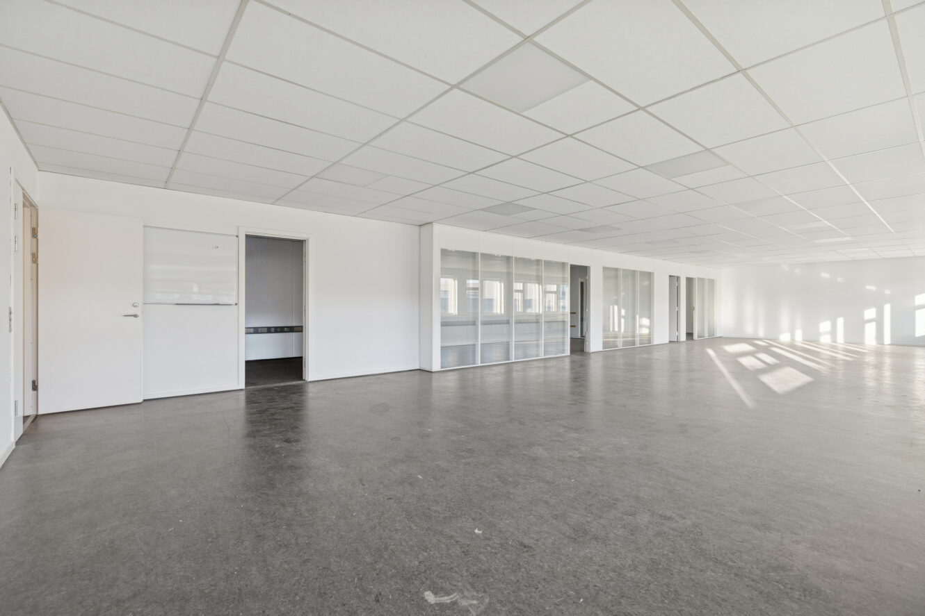 Rummeligt, tomt kontor med hvide vægge, glasskillevægge og store vinduer, der lukker naturligt lys ind.