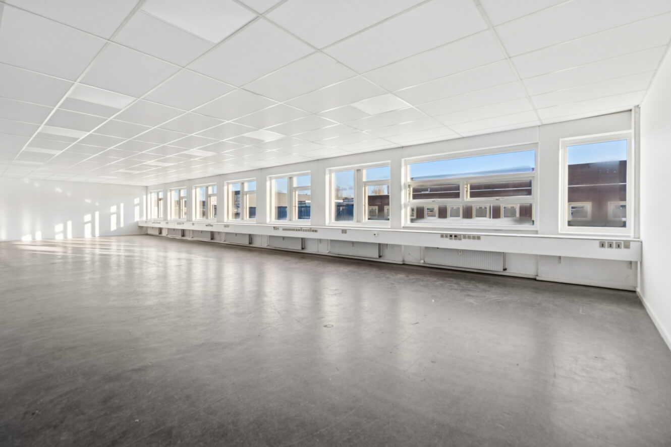 Stort, tomt kontorlokale med en række vinduer langs den ene væg, hvide vægge og loft og et bart, poleret gulv.