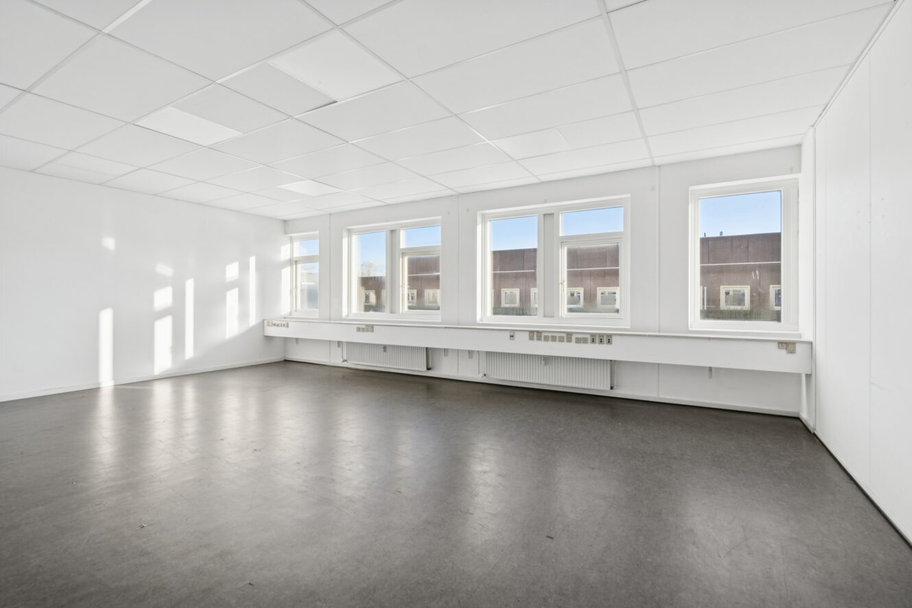 Tomt, umøbleret kontorlokale med hvide vægge, loftplader og store vinduer, der lukker naturligt lys ind; gråt gulv og udendørs bygning, der kan ses gennem vinduerne.