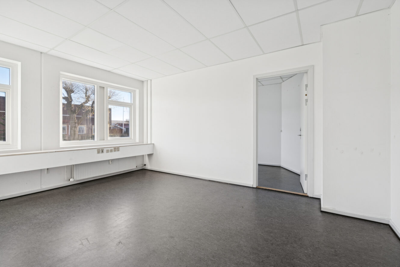 Et tomt, umøbleret rum med hvide vægge, et mørkt gulv, store vinduer på den ene side og en åben dør, der fører ind til et andet lille rum.