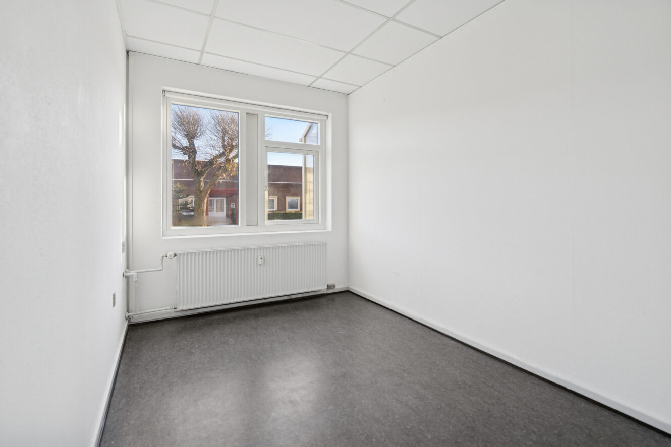 Tomt, lille rum med hvide vægge og gråt gulv, et vindue med udsigt til gaden, loftplader og en radiator under vinduet.