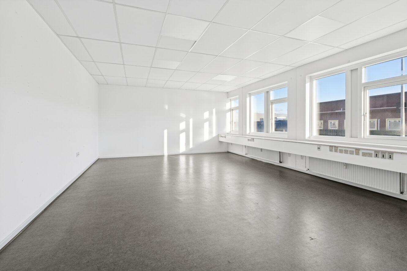 Tomt, umøbleret rum med hvide vægge, store vinduer, der giver naturligt lys, og et gråt gulv. Loftslamperne er installeret i et gittermønster.
