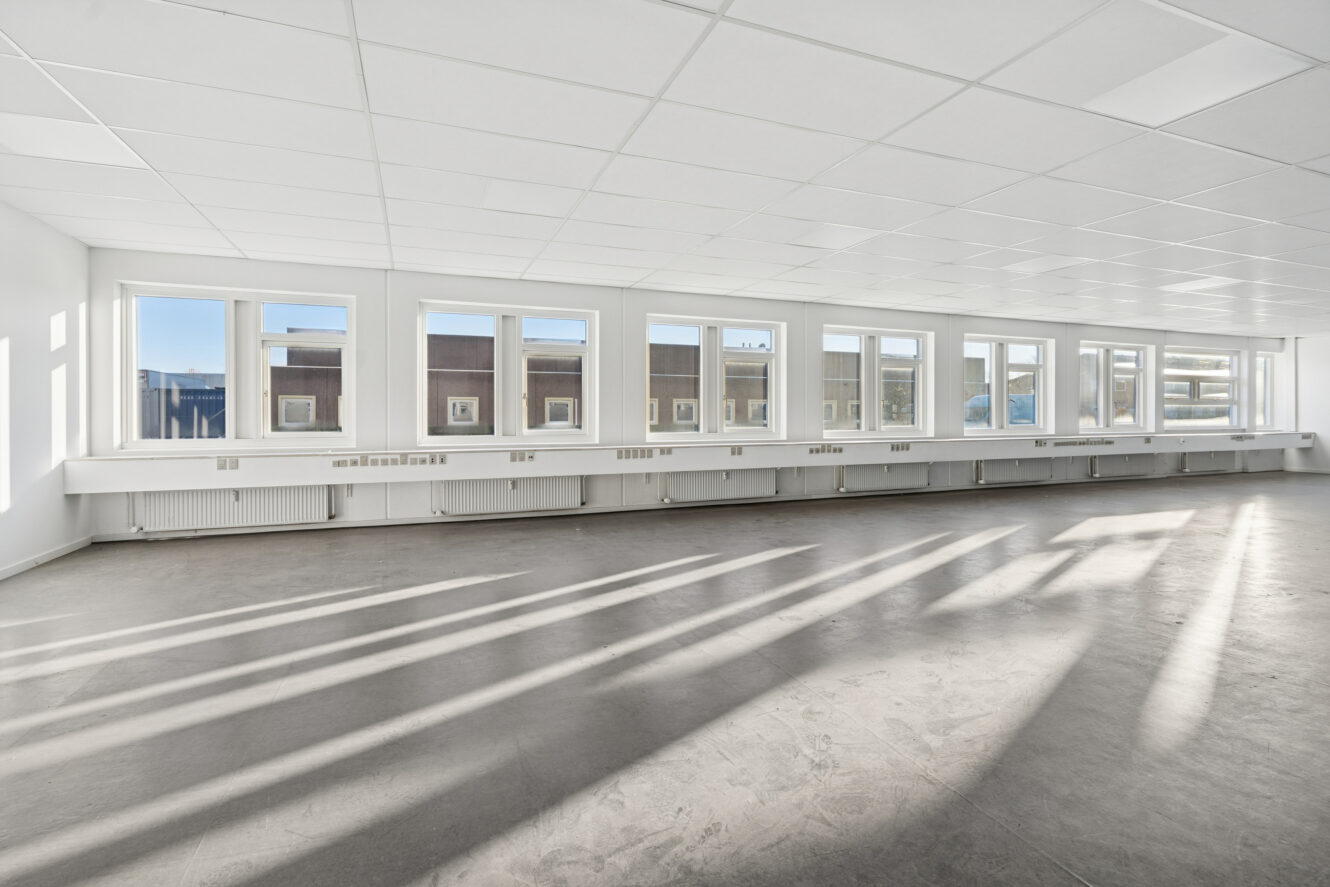 Tomt, stort rum med store vinduer, hvide vægge og et gråt gulv. Sollyset strømmer ind gennem vinduerne og kaster lange skygger hen over gulvet.