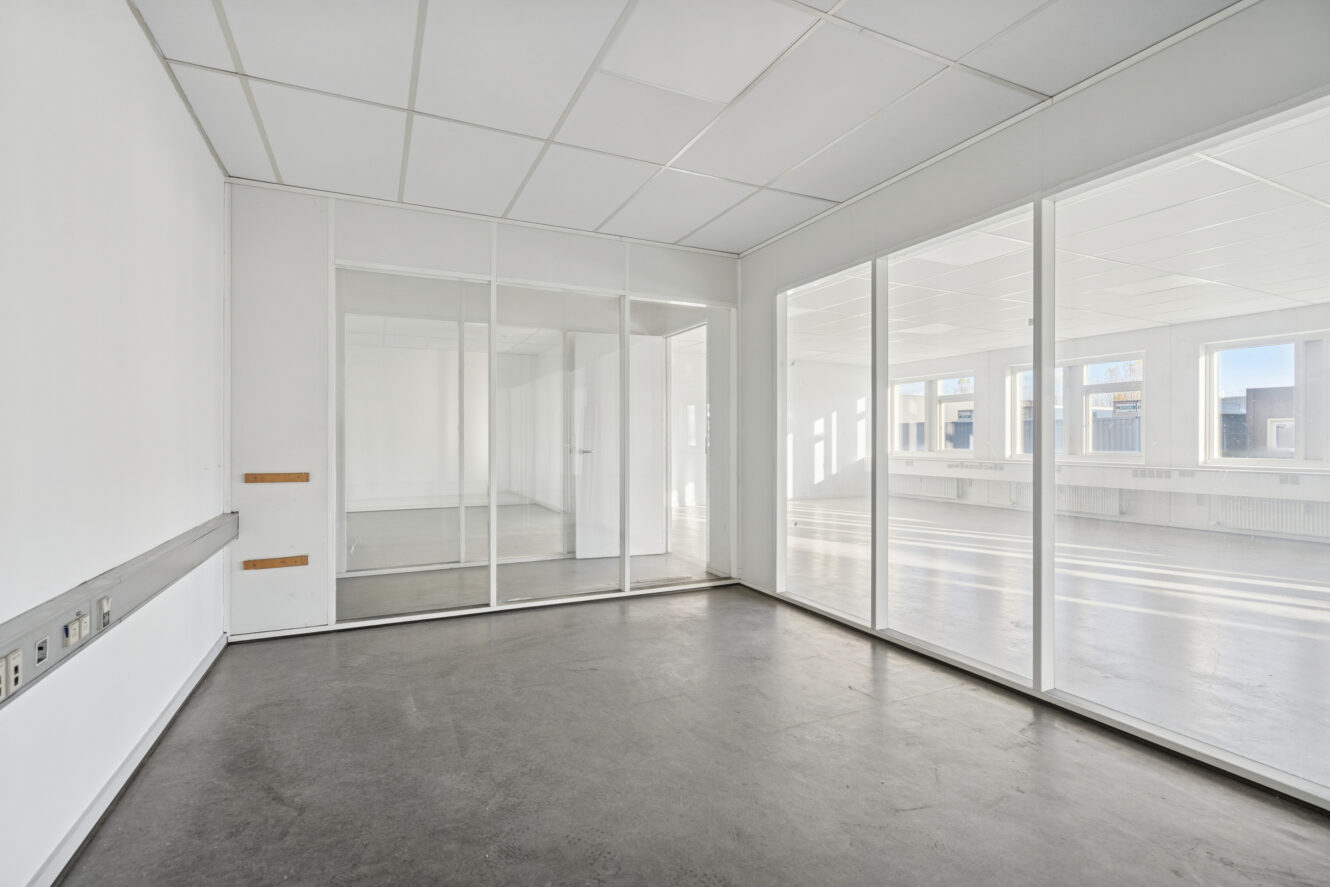 Tomt, moderne kontorlokale med hvide vægge, store glasvinduer, glasskillevæg, gråt gulv og loftplader. Stikkontakter er synlige langs den ene væg.