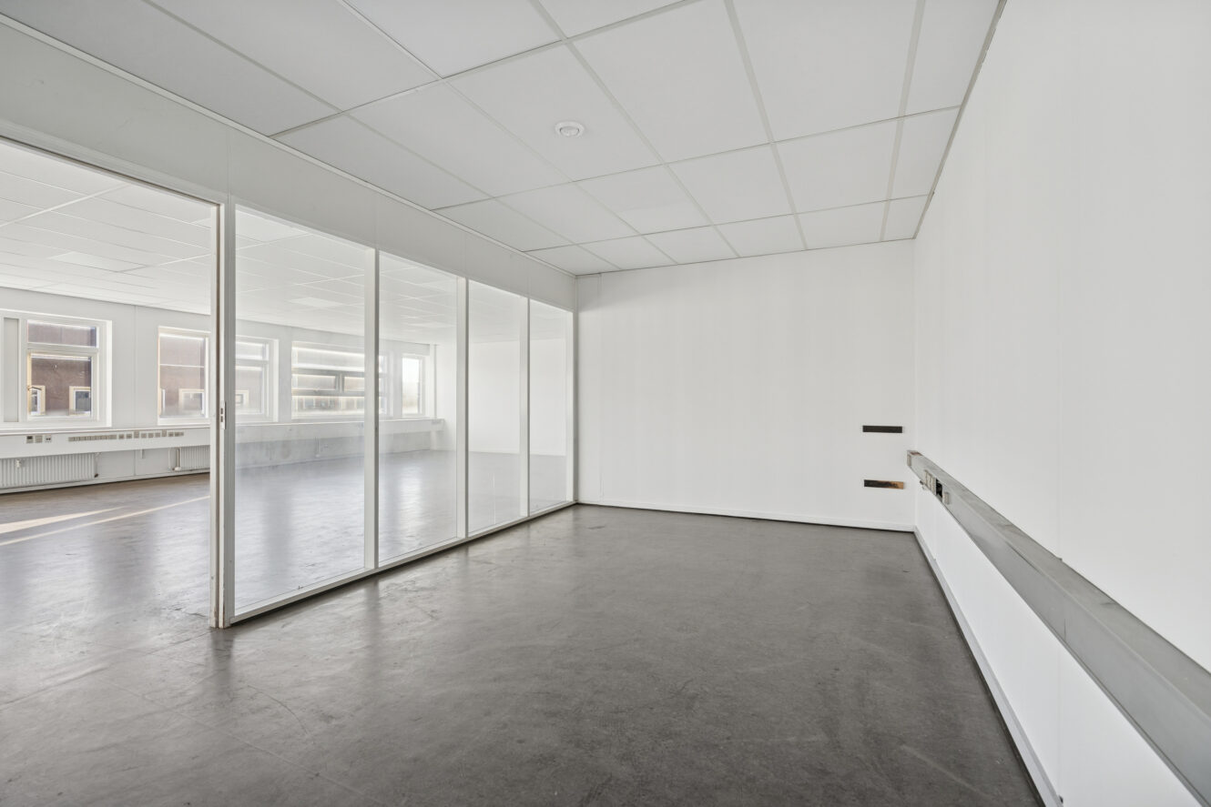 Tomt kontorlokale med hvide vægge, glasskillevæg, mørkt gulv og store vinduer, der giver naturligt lys.