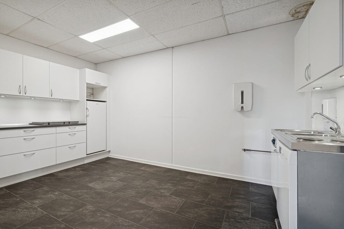 Minimalistisk køkken med hvide skabe, køleskab, vask og sort klinkegulv under et lysstofrør i et almindeligt hvidt rum.