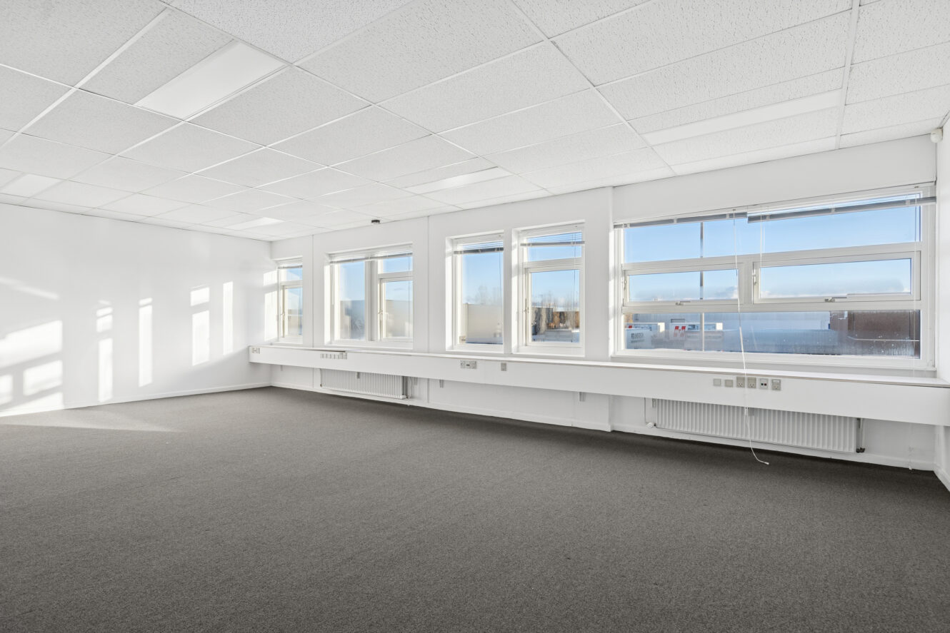 Tomme kontorlokaler med store vinduer, hvide vægge, et gråt gulvtæppe og loftslamper, der lukker naturligt dagslys ind.