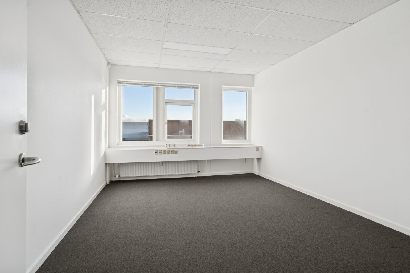 Tomt kontorlokale med hvide vægge, gråt gulvtæppe, tre vinduer og hvidt loft, set fra døråbningen.