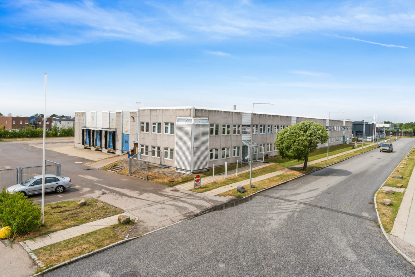 En stor, grå industriel lagerbygning med læsseramper, omgivet af en stort set tom parkeringsplads og en stille gade under en blå himmel.